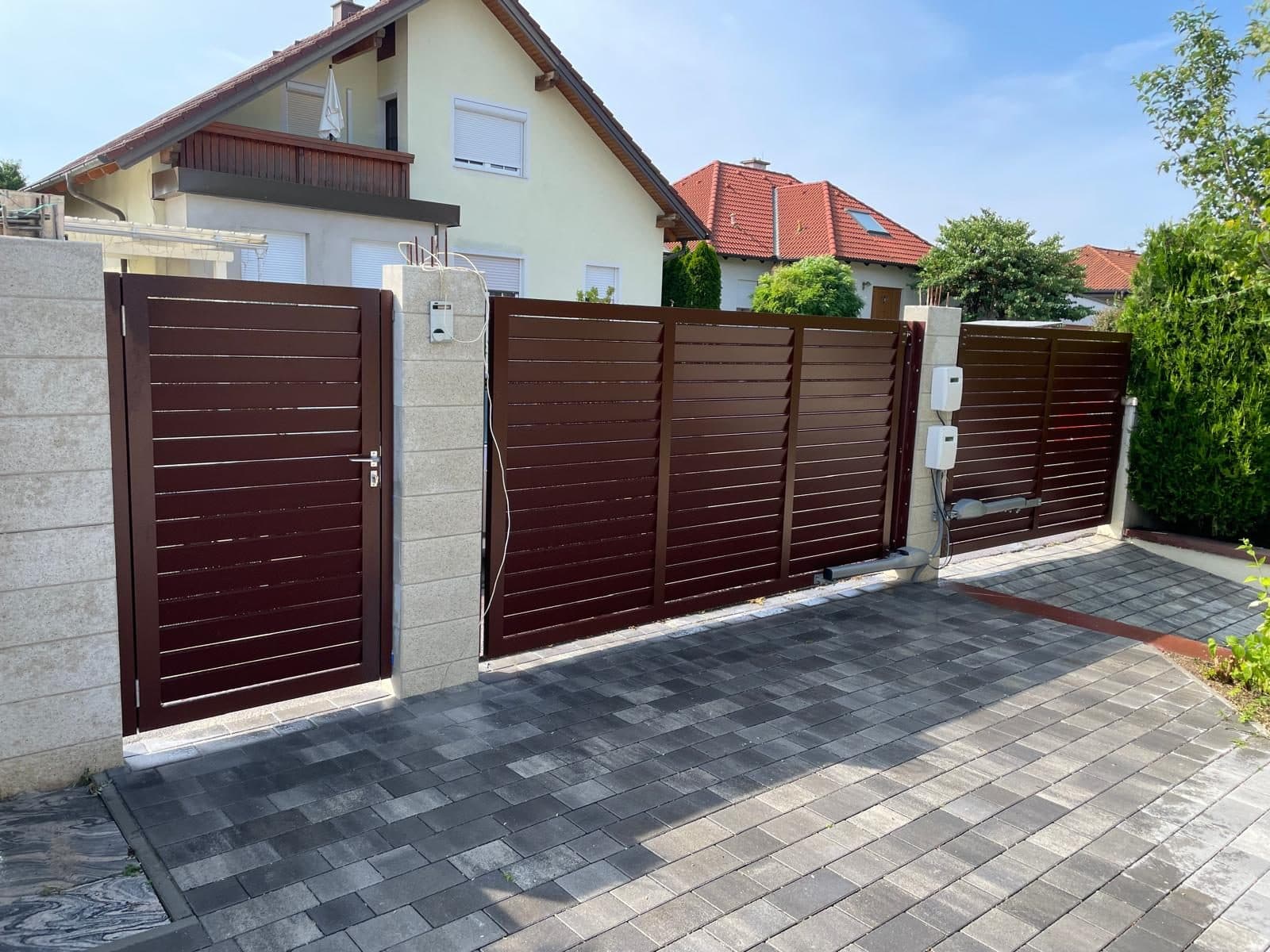 Braunes Schiebetor mit Gehtür und Steinpfeilern vor einer gepflasterten Einfahrt