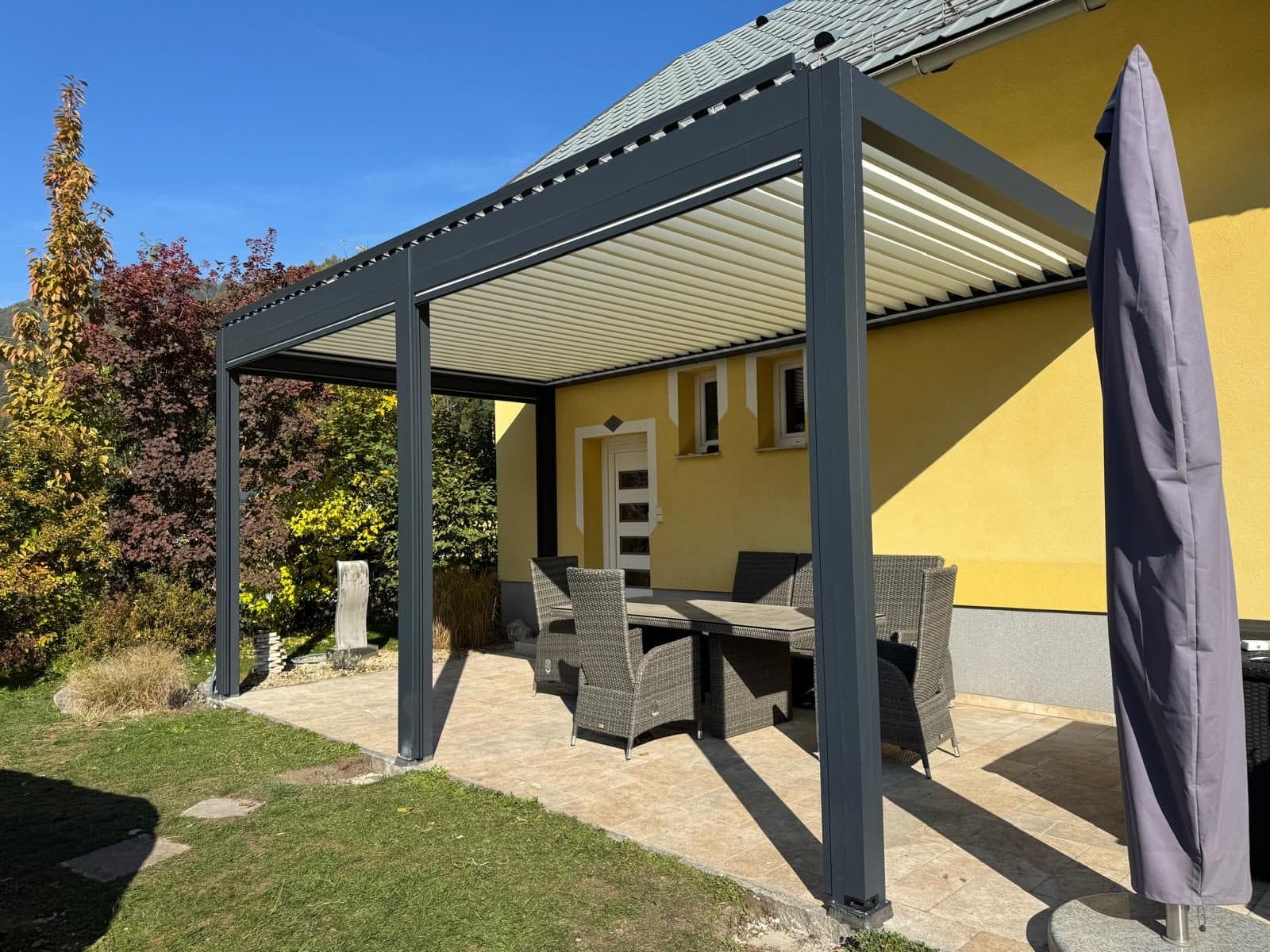 Lamellendach in Anthrazit über einer Sitzgruppe vor einem gelben Haus